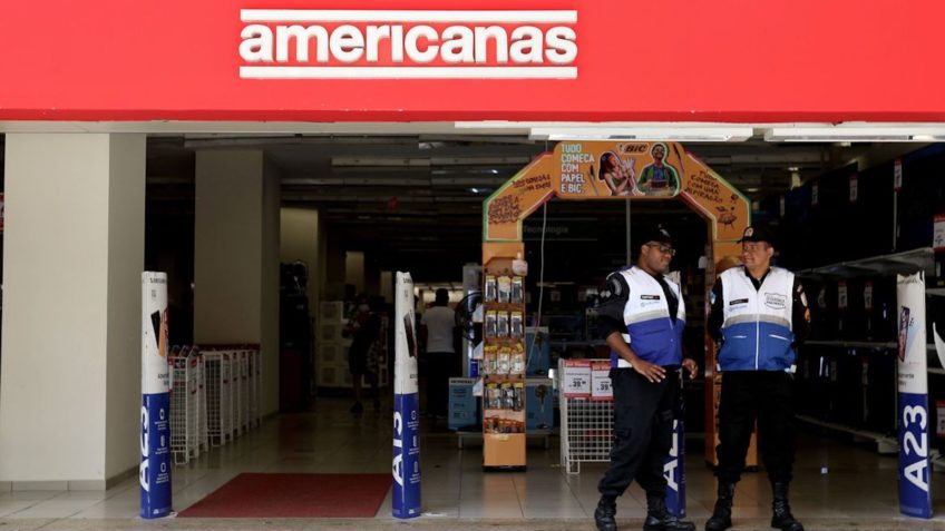Duas pessoas paradas na frente da fachada da loja das Americanas.